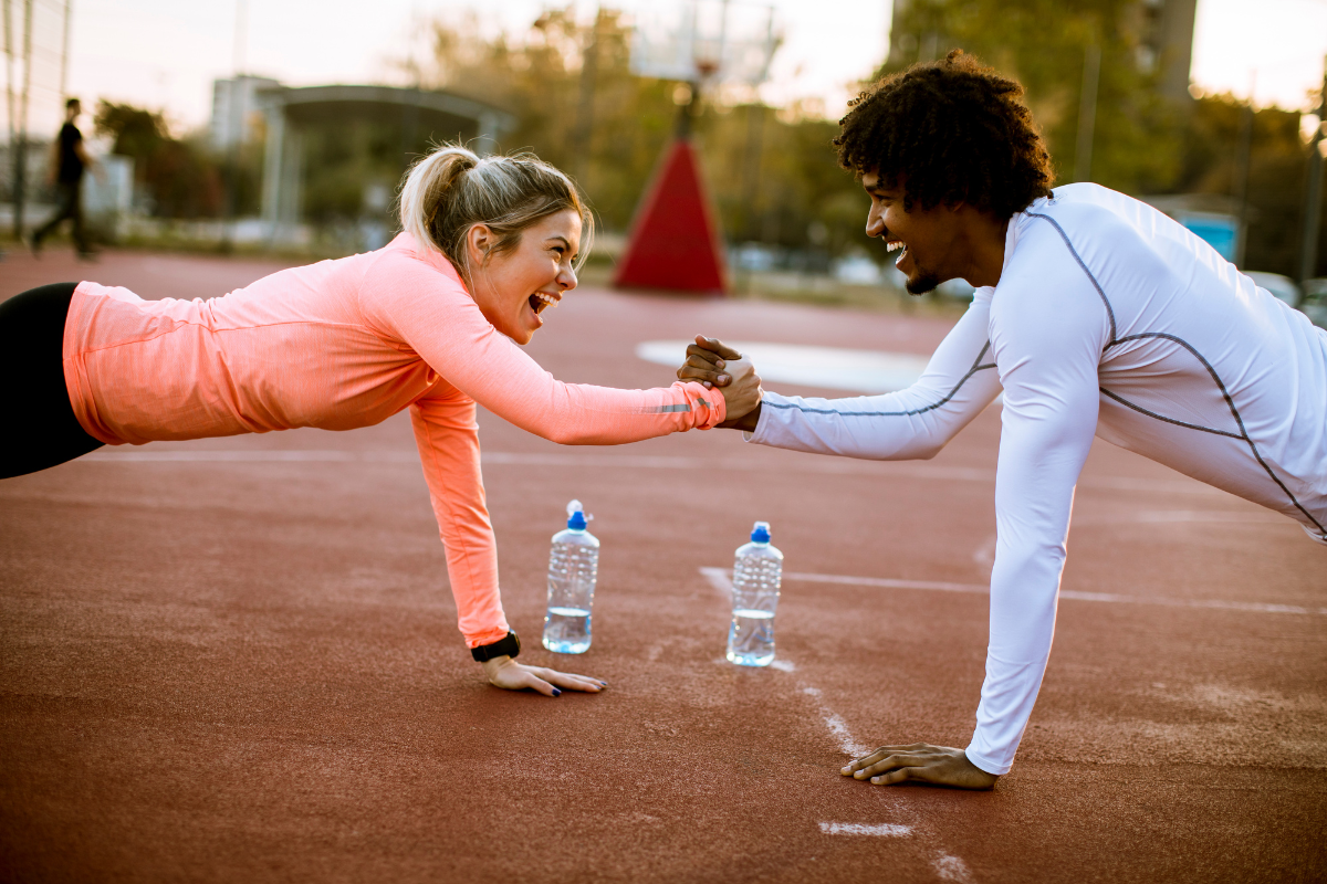 woman in peach top and man in white top in plank position gripping right hands. they are smiling and have exuberant expressions on their faces