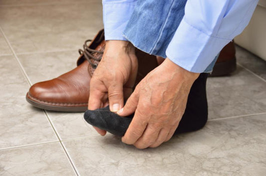 Man's hands holding a socked foot. They are wearing a light blue long sleeved shirt and there is a brown shoe in the background.