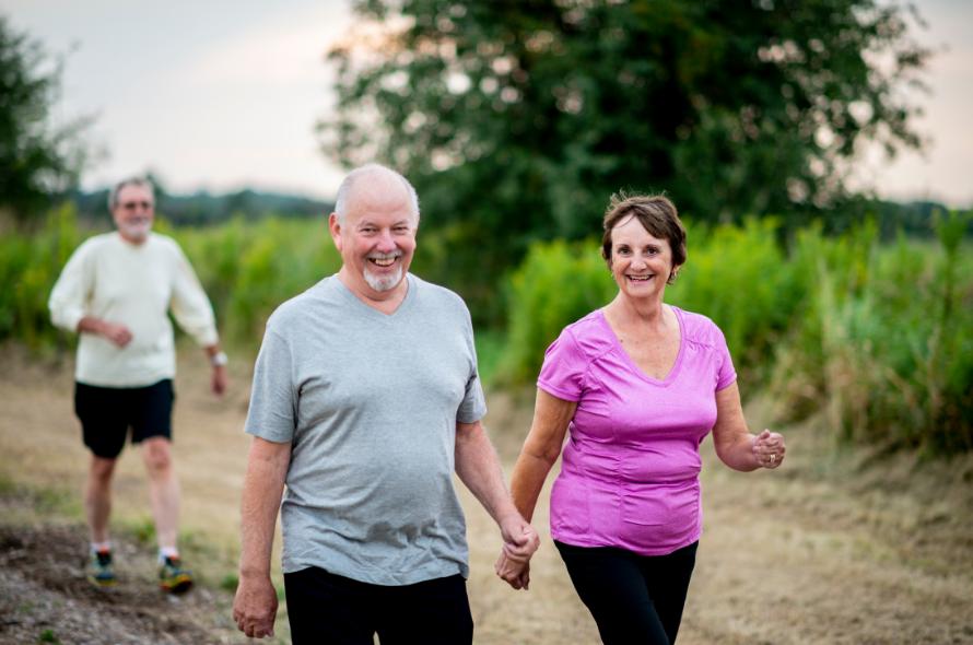 Two bearded white haired men in light tops and a woman in a pink top walking outdoors with grass and a tree in the background