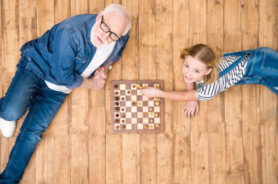 White haired man with glasses and child in pigtails and overalls lying on the floor looking up with a chessboard between them