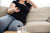 Person wearing black top and grey pants is sitting on a cream couch holding a remote. Their head and legs below the knees are out of frame.